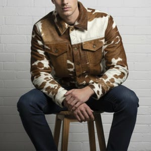 May include: A brown and white cowhide jacket with two chest pockets and button closures. The jacket has a collared neckline and is worn with blue jeans. The jacket's design features a mix of brown and white patches.
