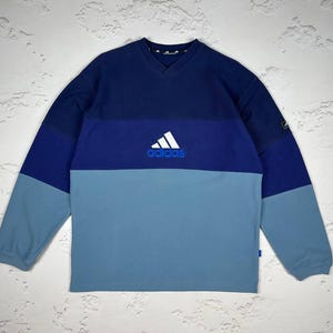 May include: A color-blocked long-sleeve sweatshirt in shades of blue. The top section is navy, the middle section is a lighter blue, and the bottom is a pale blue. The Adidas logo is embroidered in white and blue on the chest.