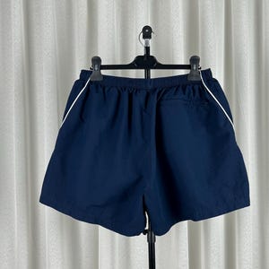 Puede incluir: Pantalones cortos deportivos azul marino con una raya blanca a lo largo de las costuras laterales. Los pantalones cortos tienen una cintura el&aacute;stica y un peque&ntilde;o bolsillo en la parte trasera. Colgados de una percha negra sobre un fondo blanco.