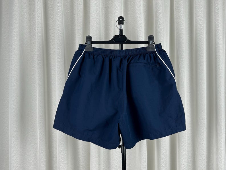 Puede incluir: Pantalones cortos deportivos azul marino con una raya blanca a lo largo de las costuras laterales. Los pantalones cortos tienen una cintura el&aacute;stica y un peque&ntilde;o bolsillo en la parte trasera. Colgados de una percha negra sobre un fondo blanco.