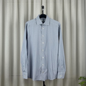 Può includere: Camicia azzurra con motivo geometrico ripetuto. La camicia a maniche lunghe è appesa a una gruccia nera su uno sfondo di tenda bianca. La camicia ha un colletto classico e polsini abbottonati.