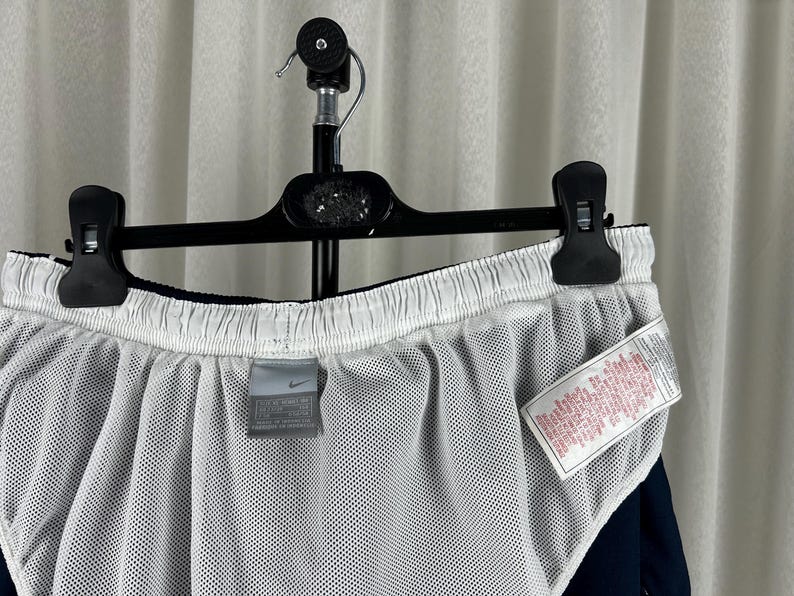 Puede incluir: Pantalones cortos deportivos azul marino con forro de malla blanco. Los pantalones cortos cuelgan de una percha negra. Se ve una peque&ntilde;a etiqueta Nike. Tambi&eacute;n hay una etiqueta blanca con texto rojo.