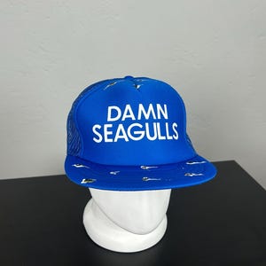 Può includere: Un cappellino trucker blu brillante con retro in rete e pannello frontale in schiuma. La scritta DAMN SEAGULLS è stampata in bianco sulla parte anteriore. La visiera piatta è decorata con un motivo ripetuto di piccoli gabbiani.