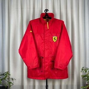 May include: A bright red Ferrari jacket with a yellow interior lining. The jacket features a yellow zipper, a drawstring waist, and the Ferrari logo on the chest. The jacket is hanging on a black hanger.