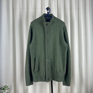 May include: A green button-up cardigan with a high collar and two front pockets. The sweater is made of a knit material and hangs on a black hanger against a white curtain backdrop.