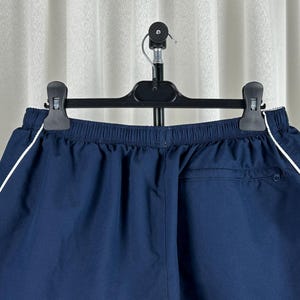 Puede incluir: Pantalones cortos deportivos azul marino con una raya blanca a lo largo de las costuras laterales. Los pantalones cortos tienen cintura el&aacute;stica y un bolsillo trasero. Cuelgan de una percha negra contra una cortina blanca.