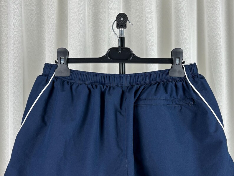 Puede incluir: Pantalones cortos deportivos azul marino con una raya blanca a lo largo de las costuras laterales. Los pantalones cortos tienen cintura el&aacute;stica y un bolsillo trasero. Cuelgan de una percha negra contra una cortina blanca.