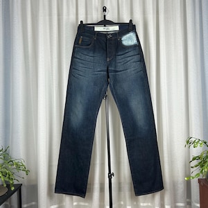 Op de afbeelding: Donkerblauwe denim jeans met een rechte pijp. De jeans worden op een hanger gepresenteerd tegen een witte gordijnachtergrond. De jeans hebben een klassiek ontwerp met vijf zakken en een knoop- en ritssluiting.