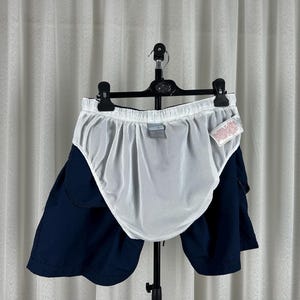Puede incluir: Pantalones cortos de ba&ntilde;o azul marino con forro de malla blanco, colgados de una percha negra. Los pantalones cortos est&aacute;n colocados frente a una cortina blanca. Una peque&ntilde;a etiqueta est&aacute; adherida a los pantalones cortos.