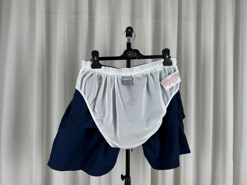 Puede incluir: Pantalones cortos de ba&ntilde;o azul marino con forro de malla blanco, colgados de una percha negra. Los pantalones cortos est&aacute;n colocados frente a una cortina blanca. Una peque&ntilde;a etiqueta est&aacute; adherida a los pantalones cortos.