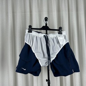Puede incluir: Pantalones cortos deportivos blancos y azul marino con cintura con cord&oacute;n. Los pantalones cortos tienen un forro de malla blanca y paneles laterales azul marino. Un peque&ntilde;o logotipo blanco de Nike es visible en la pierna izquierda.