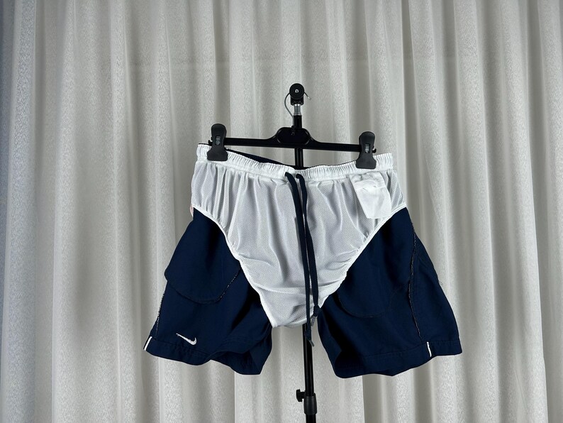 Puede incluir: Pantalones cortos deportivos blancos y azul marino con cintura con cord&oacute;n. Los pantalones cortos tienen un forro de malla blanca y paneles laterales azul marino. Un peque&ntilde;o logotipo blanco de Nike es visible en la pierna izquierda.