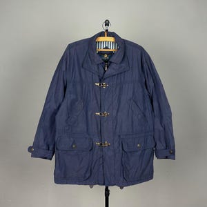 May include: A navy blue jacket featuring a fold-down collar, long sleeves with buttoned cuffs, and a front closure secured by three metal toggle clasps. The garment includes two large flap pockets at the waist and a white and blue striped lining.