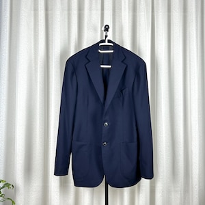 Peut inclure: Un blazer bleu marine à deux boutons, un revers cranté et deux poches avant est suspendu à un cintre blanc. La veste est présentée sur fond de rideaux blancs.