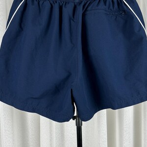 Puede incluir: Pantalones cortos deportivos azul marino con una raya blanca a lo largo de las costuras laterales. Los pantalones cortos tienen una cintura el&aacute;stica y un bolsillo trasero. La tela parece ser un material ligero y de secado r&aacute;pido, adecuado para deportes o uso casual.