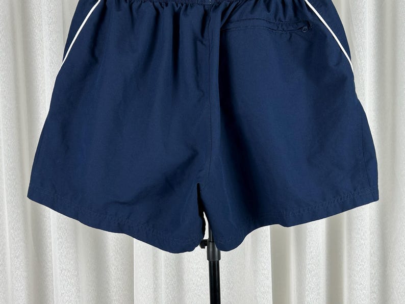 Puede incluir: Pantalones cortos deportivos azul marino con una raya blanca a lo largo de las costuras laterales. Los pantalones cortos tienen una cintura el&aacute;stica y un bolsillo trasero. La tela parece ser un material ligero y de secado r&aacute;pido, adecuado para deportes o uso casual.
