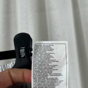 Puede incluir: Una etiqueta blanca con texto en varios idiomas, incluyendo ingl&eacute;s, franc&eacute;s y espa&ntilde;ol, que detalla la composici&oacute;n de la tela de una prenda. La etiqueta est&aacute; sujeta por una pinza negra.