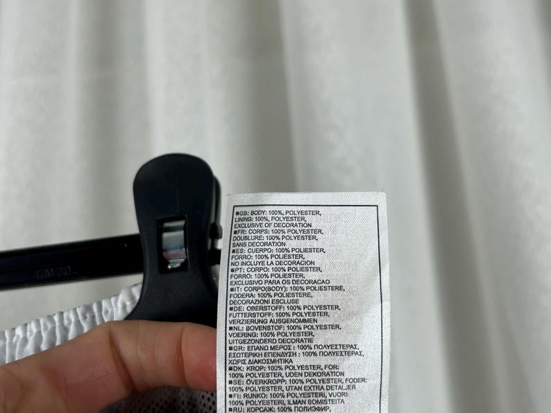 Puede incluir: Una etiqueta blanca con texto en varios idiomas, incluyendo ingl&eacute;s, franc&eacute;s y espa&ntilde;ol, que detalla la composici&oacute;n de la tela de una prenda. La etiqueta est&aacute; sujeta por una pinza negra.