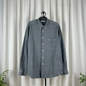 Könnte beinhalten: Ein graues Button-Down-Hemd mit langen Ärmeln, Button-Down-Kragen und Brusttasche. Das Hemd hängt an einem schwarzen Kleiderbügel vor einem weißen Vorhang. Das Hemd ist aus einem strukturierten Stoff.