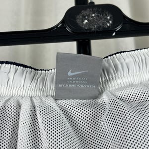Puede incluir: Primer plano de pantalones cortos deportivos blancos con forro de malla. Los pantalones cortos tienen una cintura azul marino y una etiqueta gris con el logotipo de Nike y texto. Los pantalones cortos cuelgan de una percha negra.