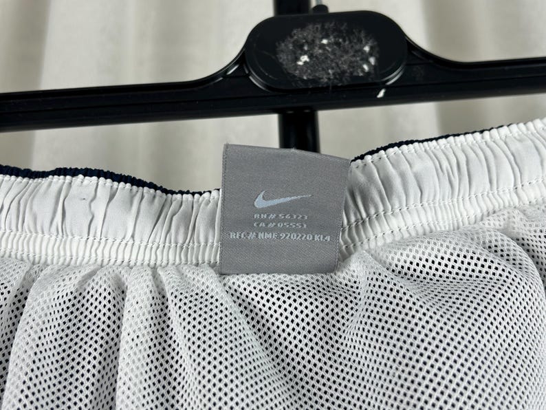 Puede incluir: Primer plano de pantalones cortos deportivos blancos con forro de malla. Los pantalones cortos tienen una cintura azul marino y una etiqueta gris con el logotipo de Nike y texto. Los pantalones cortos cuelgan de una percha negra.