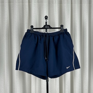 Puede incluir: Pantalones cortos deportivos azul marino con ribete blanco en los laterales. Los pantalones cortos tienen cintura con cord&oacute;n y un peque&ntilde;o logotipo blanco de Nike en la pierna izquierda. Colgados en una percha negra.