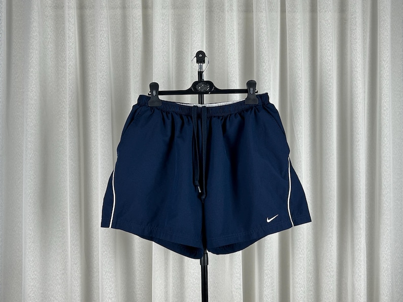 Puede incluir: Pantalones cortos deportivos azul marino con ribete blanco en los laterales. Los pantalones cortos tienen cintura con cord&oacute;n y un peque&ntilde;o logotipo blanco de Nike en la pierna izquierda. Colgados en una percha negra.