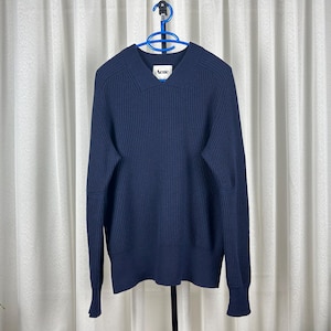 May include: A navy blue ribbed knit sweater with a collared neckline and long sleeves. The sweater is hanging on a blue hanger against a white curtain backdrop. The brand name "Acne" is visible on the collar.