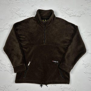 Op de afbeelding: Een bruine fleece trui met een kwart rits en een klein logo op de voorkant. De fleece heeft een wafelstructuur.