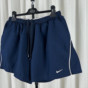 Puede incluir: Pantalones cortos deportivos azul marino con ribete blanco y cintura con cord&oacute;n. Los pantalones cortos tienen un peque&ntilde;o logotipo blanco de Nike. Cuelgan de una percha negra. Adecuados para deportes.