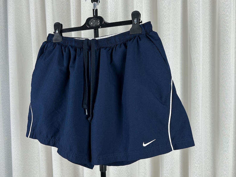 Puede incluir: Pantalones cortos deportivos azul marino con ribete blanco y cintura con cord&oacute;n. Los pantalones cortos tienen un peque&ntilde;o logotipo blanco de Nike. Cuelgan de una percha negra. Adecuados para deportes.