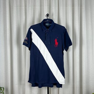May include: Navy blue polo shirt with a white diagonal stripe and a red polo player logo. The shirt has short sleeves and a collared neckline. The left sleeve features a small emblem. A classic, preppy style.