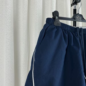 Puede incluir: Pantalones cortos azul marino con una raya blanca a lo largo de la costura lateral, colgados de una percha negra. Los pantalones cortos tienen cintura el&aacute;stica y cord&oacute;n. La tela parece ser un material ligero, adecuado para ropa informal.