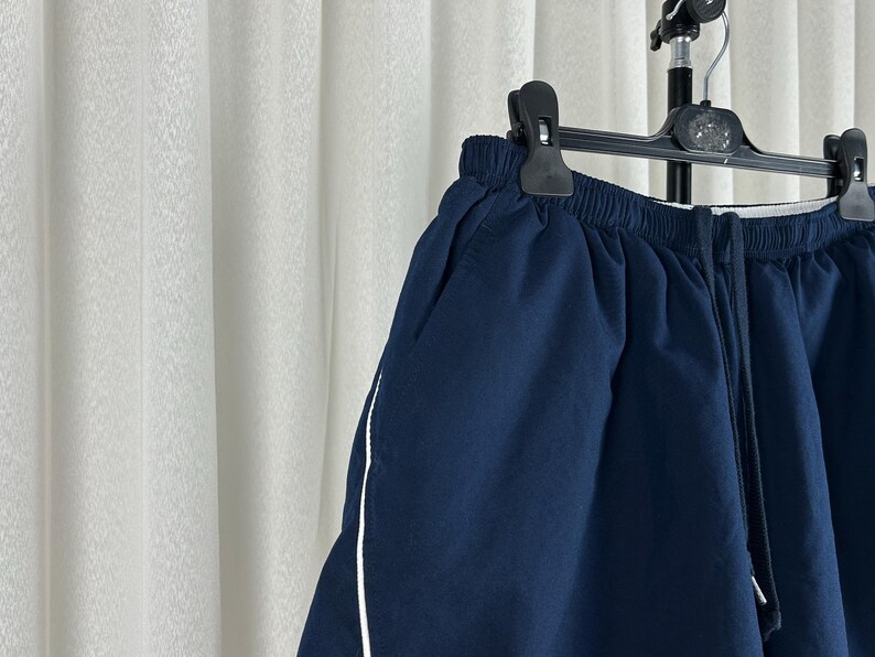 Puede incluir: Pantalones cortos azul marino con una raya blanca a lo largo de la costura lateral, colgados de una percha negra. Los pantalones cortos tienen cintura el&aacute;stica y cord&oacute;n. La tela parece ser un material ligero, adecuado para ropa informal.