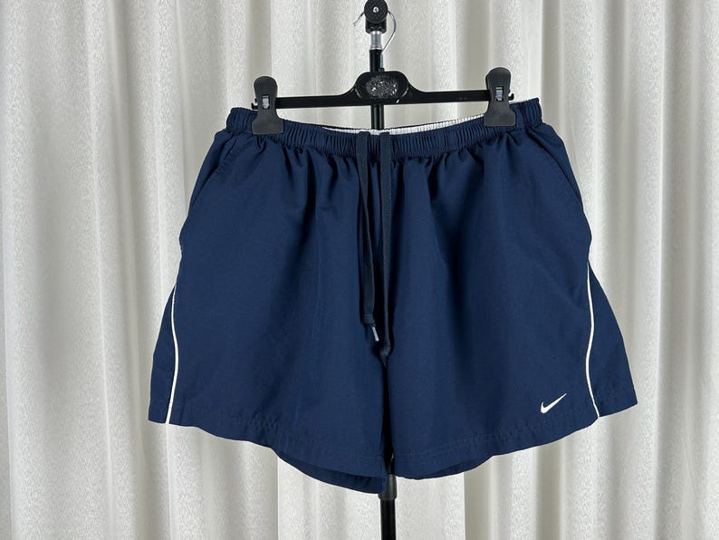 Puede incluir: Pantalones cortos deportivos azul marino con ribete blanco en los laterales. Los pantalones cortos tienen una cintura el&aacute;stica con cord&oacute;n y un peque&ntilde;o logotipo blanco de Nike en la pierna izquierda.