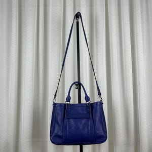 Puede incluir: Un bolso tote de cuero azul oscuro con una correa para el hombro desmontable. El bolso tiene forma rectangular con un pequeño bolsillo delantero y dos asas. El bolso cuelga de un soporte de metal negro contra un telón de fondo de cortina blanca.