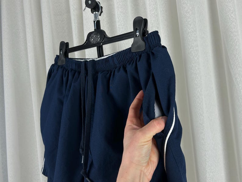 Puede incluir: Pantalones cortos deportivos azul marino con cintura con cord&oacute;n y ribete blanco en los bolsillos. Colgados de una percha negra sobre un fondo de cortina blanca. Los pantalones cortos tienen un forro de bolsillo de malla.