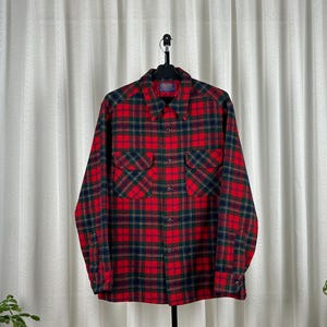 Puede incluir: Una camisa a cuadros roja, negra y verde con botones y mangas largas. La camisa tiene dos bolsillos en el pecho y está colgada de una percha negra sobre un telón de fondo blanco.
