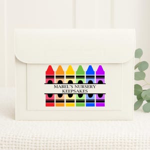 May include: A cream-coloured keepsake case with a flap closure. The front features a colourful crayon design with the text "MABEL'S NURSERY KEEPSAKES" in black. The crayons are arranged in a rainbow pattern.