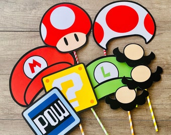 May include: A set of six colorful cardboard photo booth props featuring characters from the popular video game Super Mario Bros. The props include a red mushroom, a green question mark block, a green Luigi hat, a red and white Goomba, a black and white POW block, and a red and white Super Mario hat.