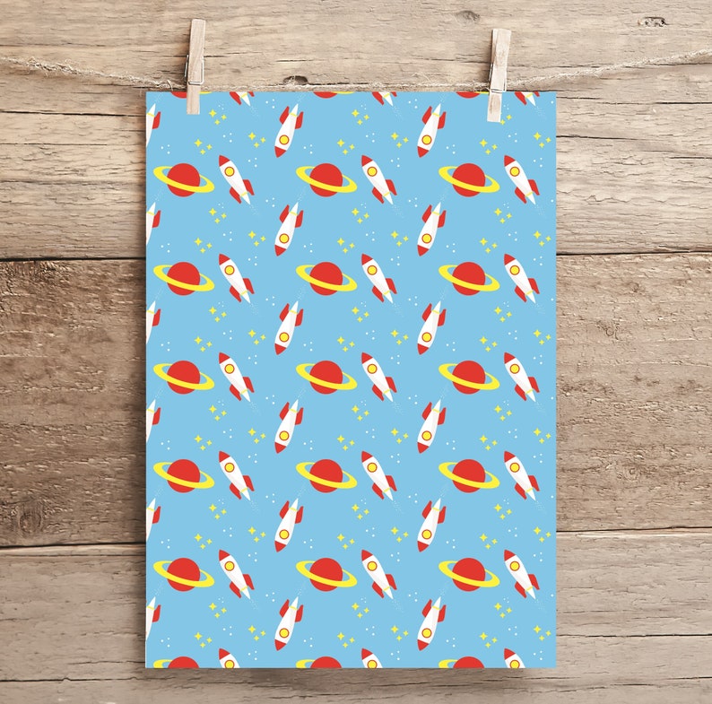 May include: A light blue paper with a repeating pattern of red and white rockets, red planets with yellow rings, and yellow stars. The paper is held by a wooden clothespin on a wooden background.