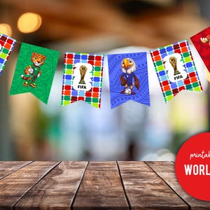 May include: A garland of pennant flags with FIFA World Cup designs. Each flag displays a cartoon mascot or the FIFA trophy. The flags are strung together and hang against a blurred background.