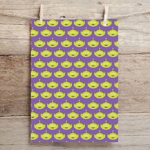 May include: A purple print featuring a repeating pattern of green, three-eyed alien characters. The print is hanging from a wooden background with a clothes pin.