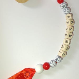 May include: A red and white baby teether with a wooden bead and a red ribbon. The teether has a soccer ball design and the letters "ARSENAL" spelled out on the wooden beads.