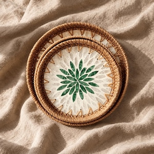 May include: Two round, woven rattan trays featuring a central inlay of white and green shell pieces arranged in a floral pattern. The trays have a natural brown finish and are stacked, showcasing the intricate weave and the decorative mosaic design.