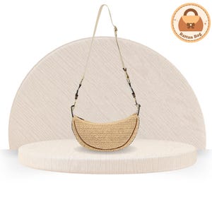 May include: A crescent-shaped rattan bag with a cream-colored strap and brown trim. The bag is displayed on a white, circular platform against a neutral background. The bag has a woven texture and a simple, elegant design.