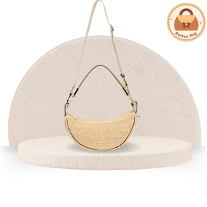 May include: A crescent-shaped rattan bag with a beige woven texture. It features a long, adjustable strap and a brown trim. The bag is displayed on a light-colored, textured surface, with a logo in the upper right corner.