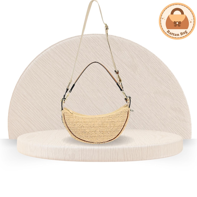May include: A crescent-shaped rattan bag with a beige woven texture. It features a long, adjustable strap and a brown trim. The bag is displayed on a light-colored, textured surface, with a logo in the upper right corner.
