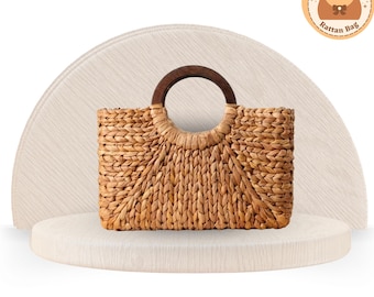 May include: A rectangular woven rattan bag with a dark brown wooden handle. The bag is light brown and has a circular handle. The bag is displayed on a white platform.