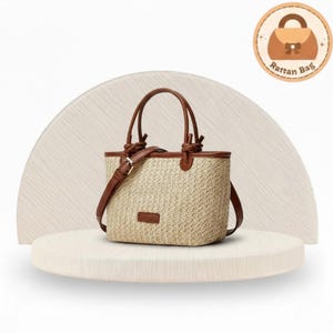 May include: A beige rattan tote bag with brown leather handles and trim. The bag has a rectangular shape with a shoulder strap and a small rectangular leather patch. The bag is displayed on a white platform with a light brown background. The text "Rattan Bag" is in a circle.